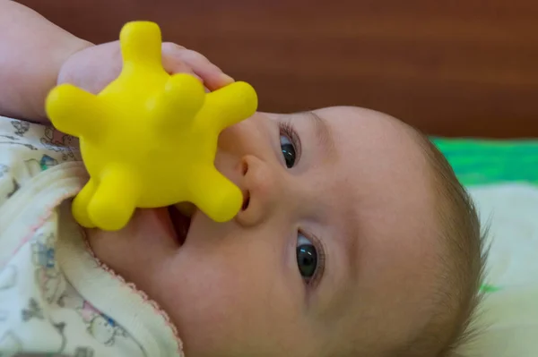 Yarım yaşındaki bir çocuk gülümsüyor ve boynuzlu sarı bir dokunsal topu ağzında tutuyor, tıpkı koronavirüs gibi.