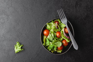Green salad leaves with cherry tomatoes in bowl on black table. Fresh and healthy food with copy space. Top view.