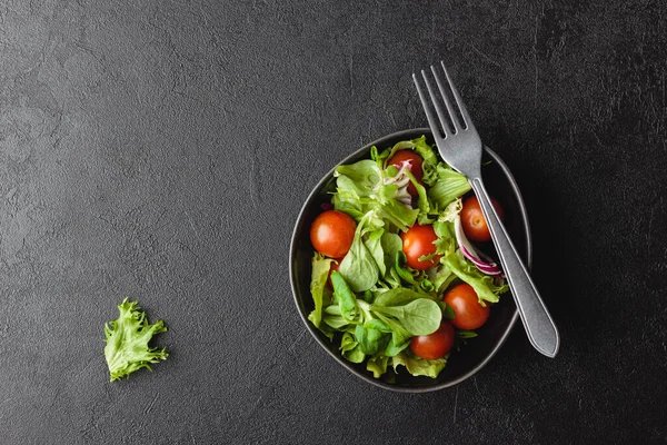 Green salad leaves with cherry tomatoes in bowl on black table. Fresh and healthy food with copy space. Top view.
