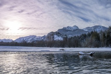 Nehirde karla kaplı alüvyonlu ağaç gövdesi. Hahnenkamm dağ sırasının önünde. Kışın gökyüzünde heyecan verici bulutlar var.