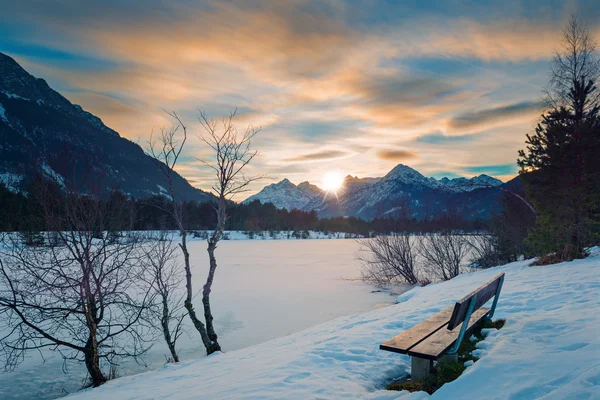 Одинокая скамейка в парке на закате в Альпах с замерзшим озером зимой — стоковое фото