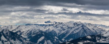 Avusturya 'nın Zell am bölgesindeki karlı dağ zirveleri. Arka planda dramatik bulutlarla dolu mavi bir gökyüzü var..