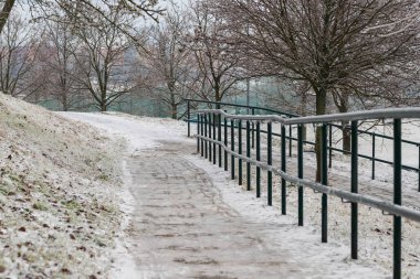 Kışın dar yol buzla kaplıdır. Yolun kenarında buzla kaplı metal bir korkuluk var. Arka planda karlı ağaçlar var..