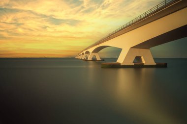 Zeeland Köprüsü, denizin üzerinde uzun beyaz bir köprü. Köprü güzel bir akşam ışığı ile aydınlatılır. Güneş tarafından aydınlatılan bulutlar. Oosterschelde Nehri 'nin karanlık yüzeyi.