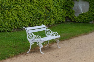 Yolda beyaz metal bir bank duruyor. Bankın arkasında bir çim ve yeşil bir duvar var..