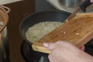 Yakından çekilen fotoğraf yemek pişirme sürecini yakalıyor, özellikle ahşap bambu kesme tahtasından kesilmiş beyaz soğanları sıcak indüksiyon tenceresine yerleştirilmiş siyah bir tavaya aktarıyor..