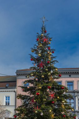 Fotoğraf, halka açık bir meydanda, açık mavi gökyüzüne karşı kurulmuş büyük ve zengin bir Noel ağacı yakalıyor..