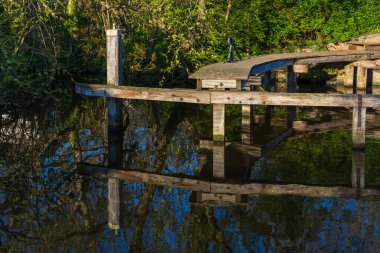 Karanlık su üzerindeki ahşap bir rıhtımın yakın çekimi, sakin yüzeye yansıyor. Bina yemyeşil bir yay tarafından kuşatılmış ve sabah güneşiyle yıkanmış..
