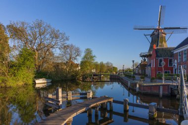Bedum 'da açık bir gökyüzünün altındaki bir kanalda yel değirmeni. Baharın yeşilliğinde ahşap bir iskele ve tekne. Klasik bir Hollanda manzarası.