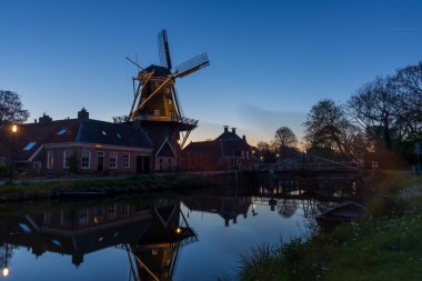Bedum 'da alacakaranlıkta aydınlatılmış bir kanalın yanındaki yel değirmeni. Geleneksel evler, bir köprü ve suda yansıyan ışıklar bir gece sahnesi yaratır..