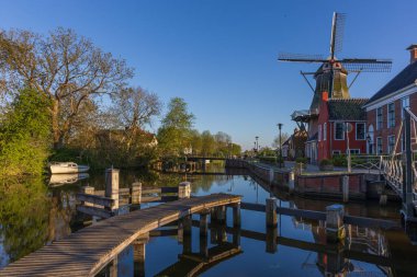 Bedum 'da yel değirmenli geleneksel Hollanda kanal sahnesi. Sakin bir bahar sabahı, berrak gökyüzü ve sudaki yansımalar, ahşap iskele ve eski binalar.-