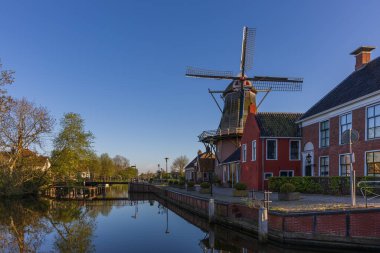 Geleneksel Hollanda yel değirmeni ve Bedum 'da kırmızı bir ev. Bahar sabahı, berrak mavi gökyüzü ve sudaki sakin yansımalar, tarihi sahne..