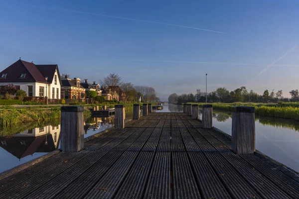 Hollanda, Bedum 'daki bir kanalın üzerindeki ahşap iskeleden sabah manzarası. Geleneksel evler sabah sisi ile sakin suya yansıyor.