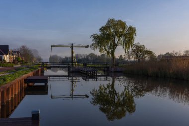 Sisli bir sabahta Bedum 'da geleneksel Hollanda asma köprüsü. Kanalda mükemmel bir yansıma, sabah bulanıklığı ve huzurlu kırsal atmosfer.