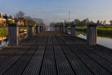 Bedum 'daki ahşap iskelenin alçak açılı perspektifi. Kanal üzerinde sabah sisi, sakin su yüzeyi ve yükselen güneşin ılık ışığı..