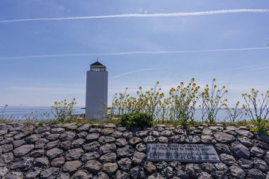 Modern beyaz deniz feneri ve Groningen 'deki deniz kıyısındaki taş bir hendekteki anıt. Bahar çiçekleri ve zıtlıklarla dolu berrak gökyüzü.
