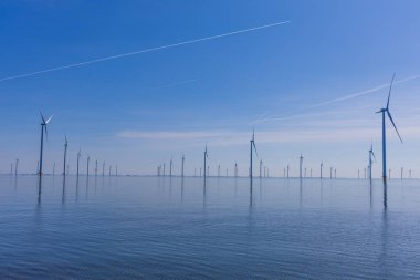 Açık mavi gökyüzünün altında durgun deniz suyunun üzerindeki modern rüzgar türbinleri. Hollanda ve Groningen eyaletlerinde sürdürülebilir enerji.