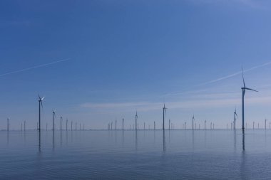 Açık mavi gökyüzünün altında durgun deniz suyunun üzerindeki modern rüzgar türbinleri. Hollanda ve Groningen eyaletlerinde sürdürülebilir enerji.