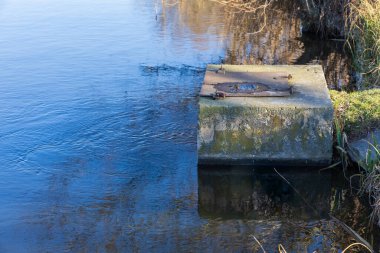 Zarostly göletindeki teknik bir su yönetim tesisinin detaylı bir görüntüsü. Yıpranmış çelik kapaklı beton yapı, kırsal altyapı ve gölet bakımını temsil eden mavi su yüzeyine yansıyor..