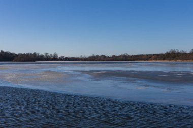 Kış sonlarında geniş bir göl ya da göl manzarası. Görüntü, açık mavi su ve ince eriyen buz tabakaları arasındaki geçişi gösteriyor. Açık mavi bir gökyüzü ve çıplak ağaçlarla dolu uzak bir kıyı şeridi sakin ve soğuk bir doğal atmosfer yaratır. P