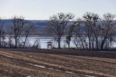 Kış kırsalının manzarası ön planda kar kalıntılarıyla dolu bir tarlanın manzarası. Ökseotuyla dolu çıplak yaprak döken ağaçlardan oluşan bir sıra donmuş bir gölün önünde duruyor. Küçük bir ahşap av standı ya da gözlem kulesi.