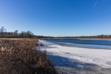 Kışın kısmen donmuş bir gölün geniş açılı manzarası. Yoğun kahverengi sazlıklar kıyı boyunca uzanır, buzlu bölgelere ve açık mavi sulara dönüşür. Gökyüzünde açık mavi gökyüzünde, bir jet uçağı uzun beyaz bir iz bırakır, pullanma hissi katar.