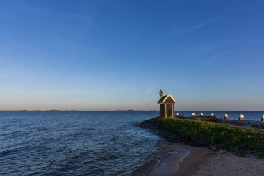 Hollanda 'daki Volendam limanının huzurlu bir akşam manzarası. Çekimde, arka planda sakin su ve yelkenli direkleri bulunan derin mavi bir alacakaranlık gökyüzünün altında aydınlık bir deniz feneri ve simgesel Mary heykeli yer alıyor..