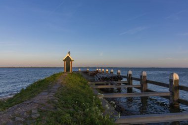 Hollanda 'daki Volendam limanının huzurlu bir akşam manzarası. Çekimde, arka planda sakin su ve yelkenli direkleri bulunan derin mavi bir alacakaranlık gökyüzünün altında aydınlık bir deniz feneri ve simgesel Mary heykeli yer alıyor..