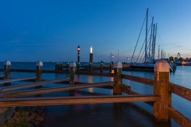 Hollanda 'daki Volendam limanının huzurlu bir akşam manzarası. Çekimde, arka planda sakin su ve yelkenli direkleri bulunan derin mavi bir alacakaranlık gökyüzünün altında aydınlık bir deniz feneri ve simgesel Mary heykeli yer alıyor..