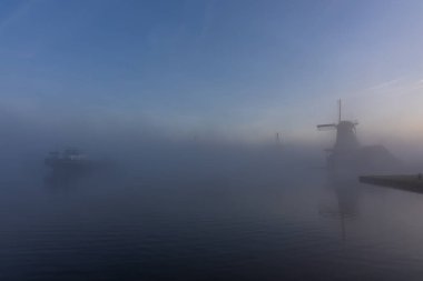 Hollanda 'dan çekilen ve içinde bir tekne ve yoğun sislerin içinde kaybolan tarihi bir yel değirmeninin yer aldığı atmosferik bir sabah çekimi. Sakin su yüzeyi, şafağın yumuşak mavi ışığını yansıtır. Büyük bir kopyayla minimalist ve dingin bir bileşim oluşturur.