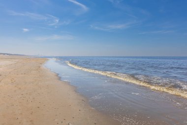 Hollanda 'daki Kuzey Denizi' nin uçsuz bucaksız kumlu kıyı şeridi boyunca geniş bir manzara. Yumuşak köpüklü uzun gelgit çizgisi, uzak bir şehrin ufuk çizgisinin görülebildiği ufka doğru gözünü açar. Mavi gökyüzü ve dağınık sirrus bulutuyla aydınlık bir gün.