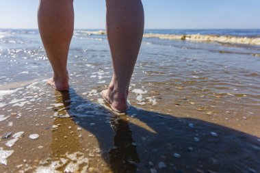 Wassenaarseslag plajında Kuzey Denizi 'nin kıyısında yürüyen birinin arka görüntüsü. Su ayak bileklerini yıkar ve ıslak kumda yumuşak köpük oluşturur. Hollanda 'da güneşli bir gün özgürlük, rahatlama ve yaz tatilini çağrıştırıyor..