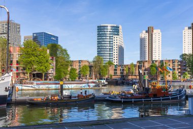 Limana demirlemiş tarihi römork ve gemilerin yer aldığı Rotterdam 'ın Leuvehaven manzarası. Modern konut ve ofis kuleleri açık mavi gökyüzünün altında arka planda yükseliyor. Bu resim Rotterdam ve kanadının dinamik gelişimini gösteriyor.