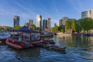 Rotterdam 'ın Leuvehaven limanındaki RB tekne deneyimi üssünün manzarası. Arka planda açık mavi gökyüzü altında modern gökdelenler ve yerleşim kuleleri yer alıyor. Görüntü turizm faaliyetlerini ve çağdaş Hollanda mimarisini yansıtıyor