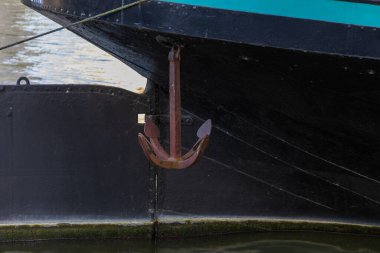Eski ve yıpranmış bir çapanın yakın plan çekimi. Siyah bir teknenin yanında görünür bir pas asılı. Arka planda sakin su görülebilir. Bu görüntü deniz tarihini, denizcilik geleneğini ve endüstriyel estetiği çağrıştırıyor. Nakliye için ideal, tra