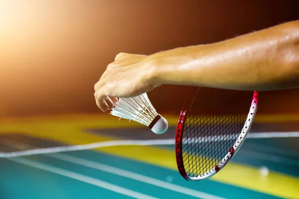 Badminton player is holding white badminton shuttlecock and badminton racket in front of the net before serving it over the net to another side of badminton court. Selective focus on white shuttlecock