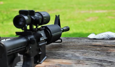 Eğitim alanındaki ahşap masada dürbünü olan uzun namlulu siyah otomatik tüfek, ayrıca Air Soft veya BB Gun olarak da bilinir, hafif ve seçici bir şekilde silah namlusuna odaklanmış..