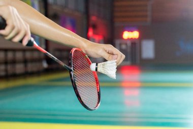 Badminton 'da düşük bir servisin ilk anlarını yakalıyoruz. Oyuncuların elleri, pembe bir raketin yanında beyaz servis aletini sabitliyor ve canlı yeşil kortta kırmızı bir çerçeve..