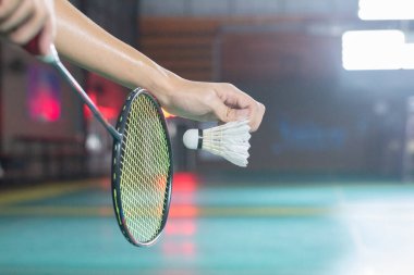 Mekik horozunun oyuncunun elinden tutulduğu güçlü bir görüntü, servis için hazır, koyu renkli raket, kortun önünde parlak sarı teller var..