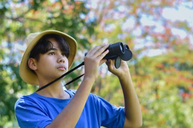 Mavi tişört ve kamuflaj şapkası takan genç bir çocuk kameradan uzak durmak için bir dürbün kullanıyor. Ormanda ya da parkta yoğun bir yeşilliğin ortasında duruyor..