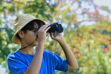 Mavi tişört ve kamuflaj şapkası takan genç bir çocuk kameradan uzak durmak için bir dürbün kullanıyor. Ormanda ya da parkta yoğun bir yeşilliğin ortasında duruyor..