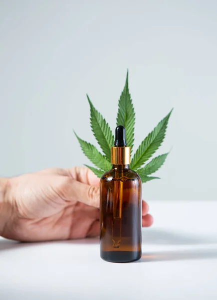 Zbliżenie słoika szklanego z produktem CBD serum cannabis i liściem konopi w dłoni na szarym tle — Zdjęcie stockowe