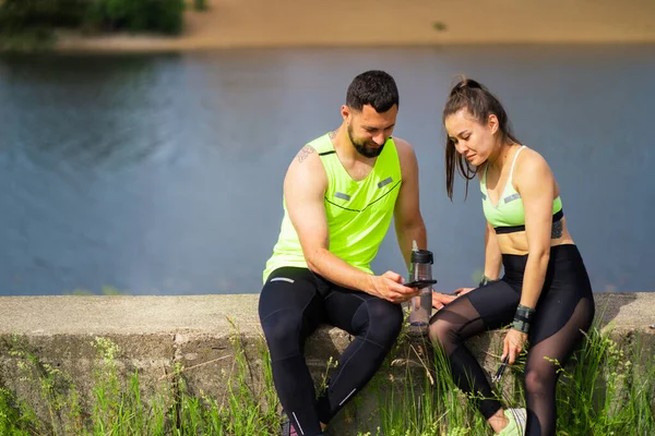 Sporty young couple in stylish sportswear looking at phone after workout out on river background — стоковое фото