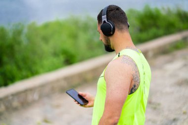 Sakallı, kulaklıklı sporcu ve şık spor kıyafetleri giyen, cep telefonunda sörf yapan spor uygulaması kullanan.