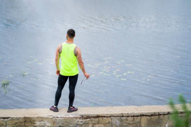 Şık spor kıyafetleri giyen atletik bir adam su arka planında bir şişeyle dikiliyor.