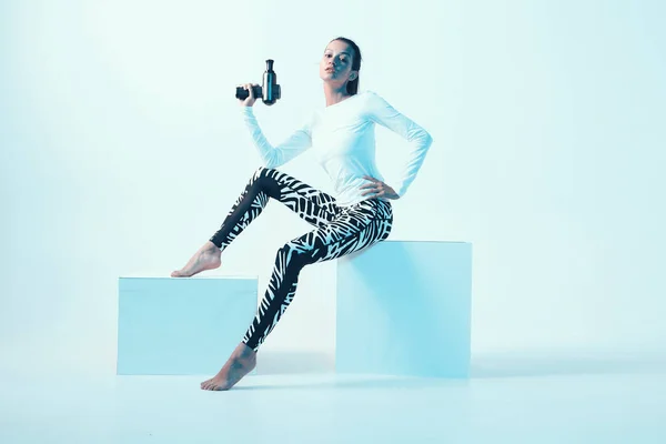 Ударный массажер, спортивная молодая женщина с ручным массажным пистолетом, сидящая на кубе в неоновом студийном свете — стоковое фото