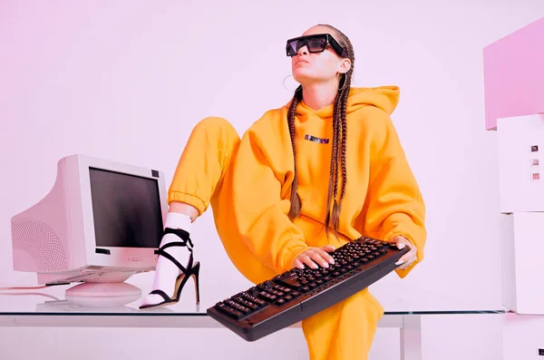 Cool young office female worker in stylish sunglasses, hoodie sitting with old computer and keyboard. Colored Neon light