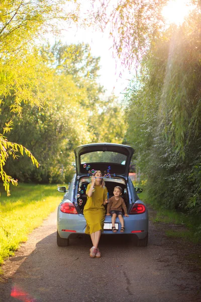 Bela mãe e pequeno filho viajantes sentar no porta-malas do carro na estrada e soprar bolhas de sabão. Foto sobre viagens em família — Fotografia de Stock