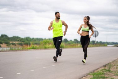 Şık spor kıyafetleriyle koşan genç bir çift. Bulutlu havada dışarıda spor yapıyorlar. Sağlıklı aktif yaşam tarzları var.