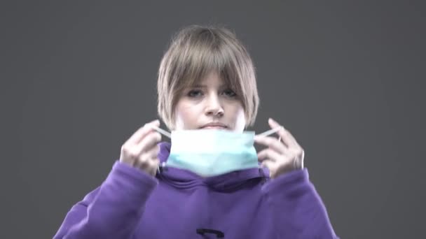 Serious young girl puts on protective medical facemask on gray background. Stop corona virus pandemic outbreak concept — Stock Video